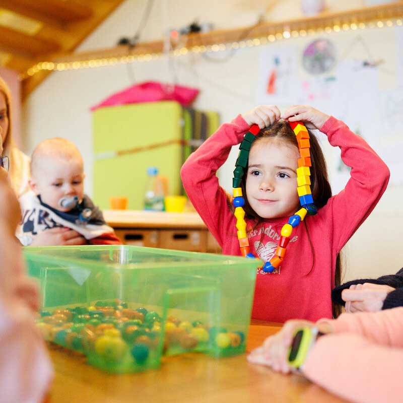 Ein junges Mädchen sitzt an einem Tisch und zieht sich bunte Perlenketten über den Kopf. Daneben sitzt ein Baby mit einem Schnuller. Auf dem Tisch stehen grüne Behälter, die mit Perlen gefüllt sind. Die Hände anderer Kinder sind zu sehen.