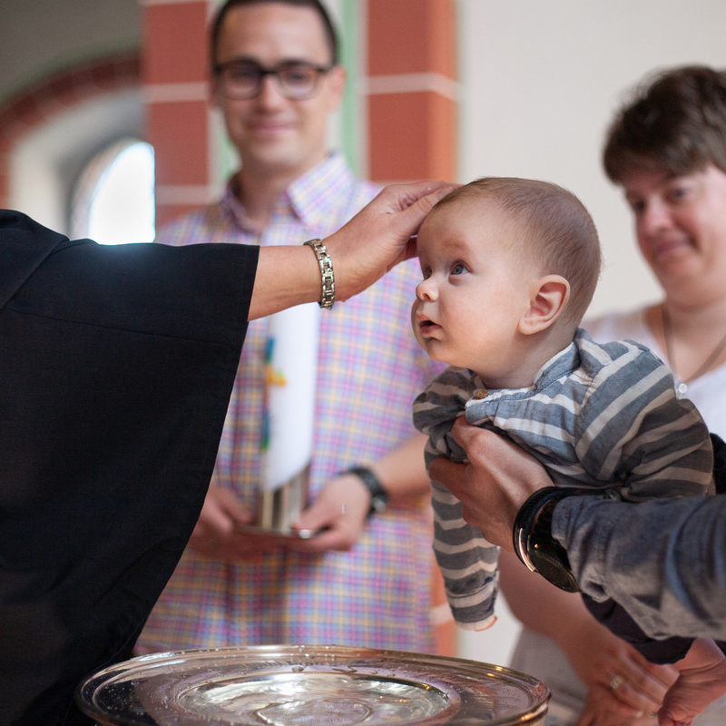 Ein Baby in gestreifter Kleidung wird über ein Taufbecken gehalten, während eine Person in einer schwarzen Robe sanft den Kopf des Babys berührt. Im Hintergrund stehen zwei Erwachsene, die die Zeremonie beobachten und lächeln.