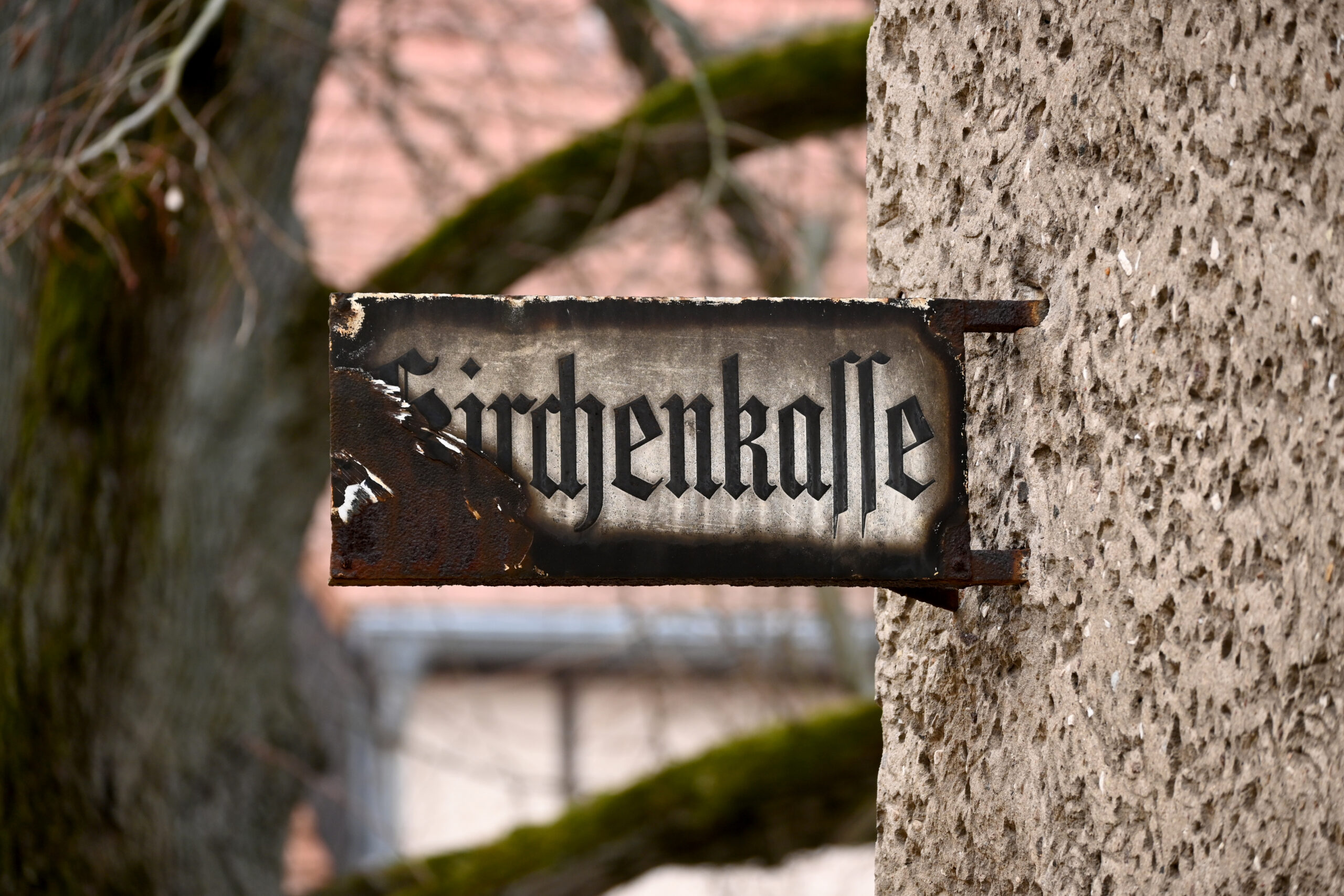 An einer rauen, beigen Wand ist ein verwittertes Metallschild mit der Aufschrift „Kirchenkasse“ in gotischer Schrift angebracht. Im Hintergrund sind Äste und ein verschwommenes Gebäude zu sehen.