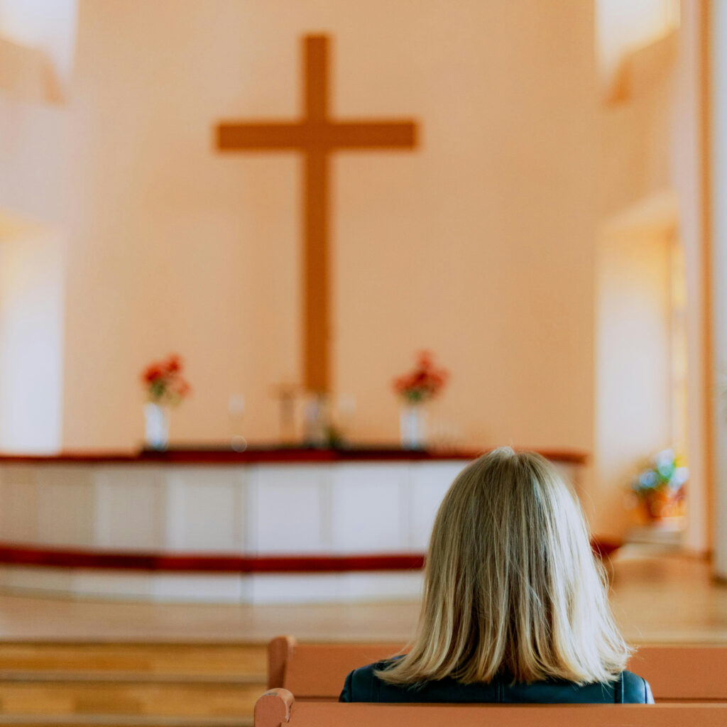 Eine Person mit blonden Haaren sitzt allein in einer Kirchenbank und blickt auf ein großes Holzkreuz am mit Blumen geschmückten Altar.