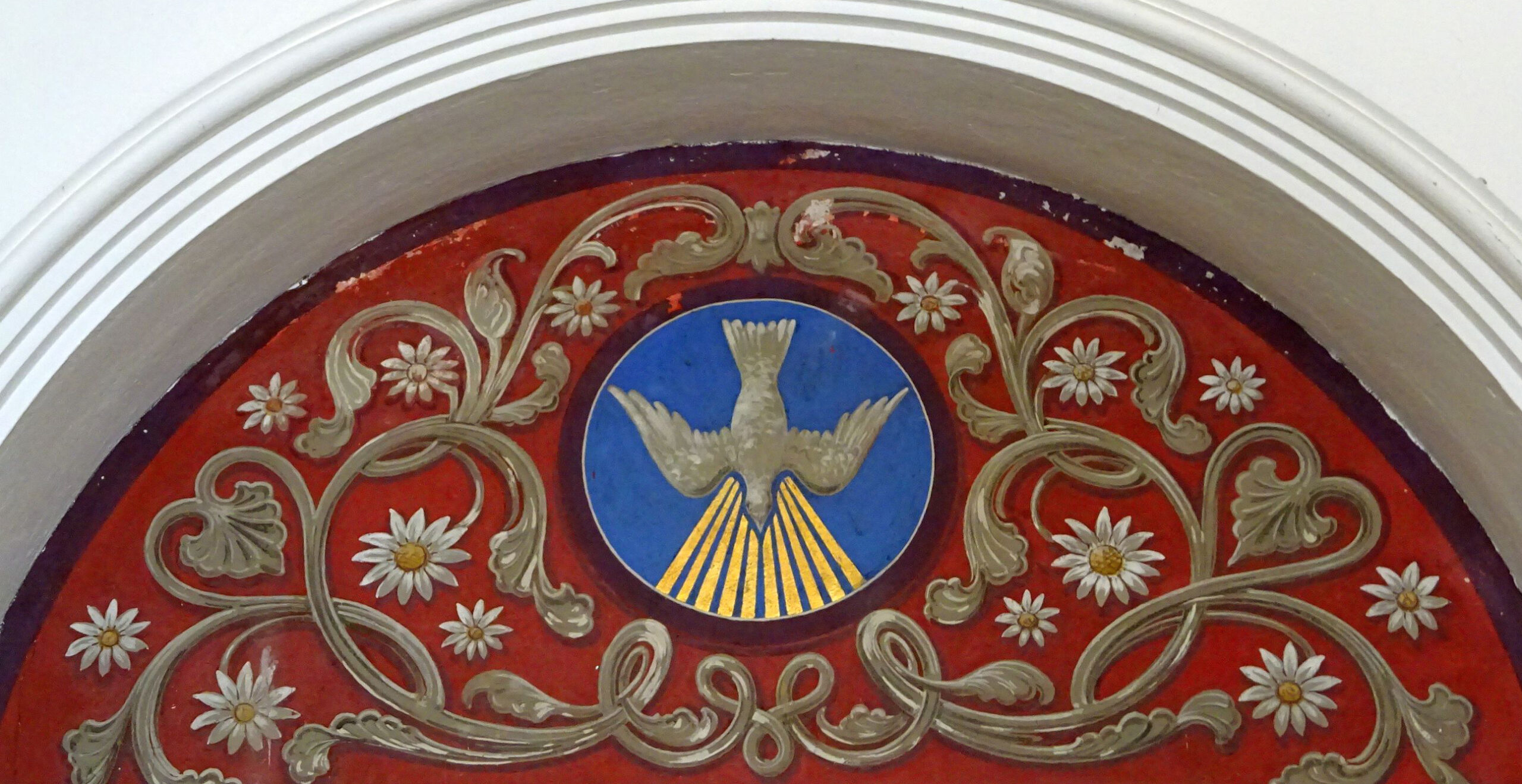 Ein dekoratives Fresko zeigt eine weiße Taube auf einem blauen Kreis mit goldenen Strahlen, umgeben von Blumenmustern auf rotem Hintergrund in einem gewölbten Rahmen.
